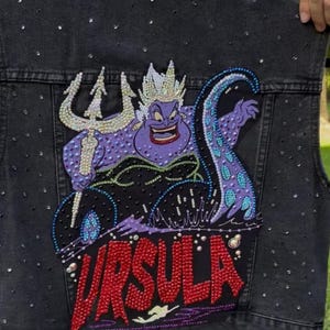 May include: Black denim vest with a bedazzled Ursula design. The design features the sea witch with a trident, purple tentacles, and the name "URSULA" in red. The vest is embellished with scattered rhinestones.
