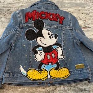 May include: A blue denim jacket with a rhinestone embellished Mickey Mouse design on the back. The design includes the word "Mickey" in red rhinestones above the character.