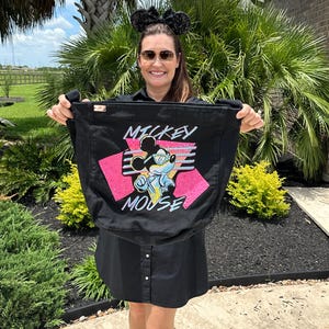 May include: Black tote bag featuring a graphic of Mickey Mouse in a retro style. The design includes the words "Mickey Mouse" in a stylized font, with pink and blue accents. The bag is held up in front of a person.
