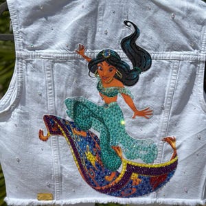 May include: White denim vest with a rhinestone embellished design of Princess Jasmine riding on a magic carpet.