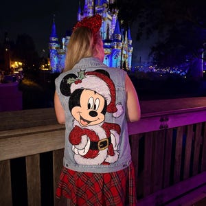 May include: A denim vest featuring a sparkling Mickey Mouse in a Santa Claus outfit. The vest is embellished with red and silver sequins. The person is wearing a red plaid skirt and a red sequined bow. The background shows a lit-up castle at night.