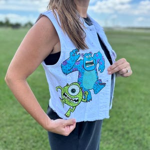 May include: A light blue denim vest featuring a colorful graphic of two cartoon monsters. The vest has a frayed hem and is embellished with small, sparkling accents. The design includes a large, blue monster and a small, green monster.