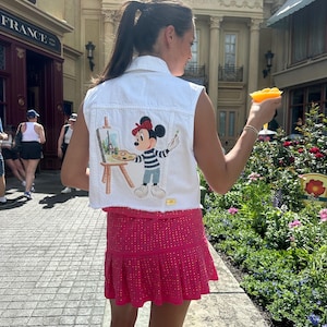 May include: A white denim vest with a Mickey Mouse painting on the back. Mickey is wearing a striped shirt and beret and is painting a picture of the Eiffel Tower. The vest has a frayed hem.
