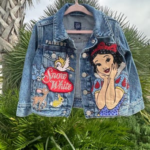 May include: A blue denim jacket with a Snow White design. The jacket has a red heart with the words "Fairest Snow White" on it. The jacket also has a design of Snow White with a bird and a deer.