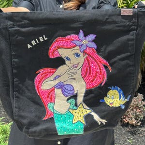 Op de afbeelding: Zwarte tote bag met een kleurrijke illustratie van Ariel uit De Kleine Zeemeermin, met roze haar, een paarse schelpenbeha en een groene staart. Het woord "ARIEL" is in witte letters geschreven. Een geel-blauwe vis is ook afgebeeld.