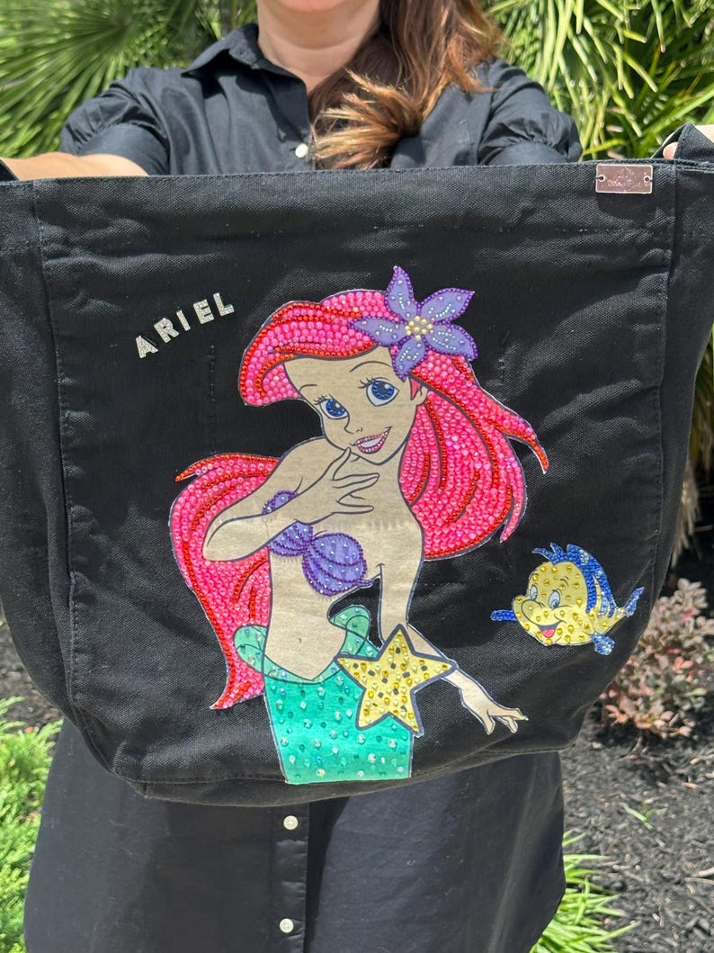 Op de afbeelding: Zwarte tote bag met een kleurrijke illustratie van Ariel uit De Kleine Zeemeermin, met roze haar, een paarse schelpenbeha en een groene staart. Het woord "ARIEL" is in witte letters geschreven. Een geel-blauwe vis is ook afgebeeld.