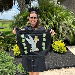 May include: Black tote bag featuring a sparkling image of Tinkerbell with the word "TINKERBELL" in green and white letters. The bag has a button closure and black handles. The bag is held by a person wearing a black dress and sunglasses.