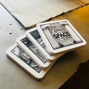 May include: Set of five white square coasters with black ink illustrations. The top coaster features a wave with the text "God fills the space he creates".