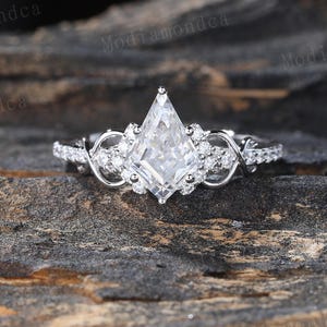 May include: A silver engagement ring with a large, kite-shaped central stone. The ring features smaller accent stones and intricate detailing along the band. The ring is set against a dark, textured background.