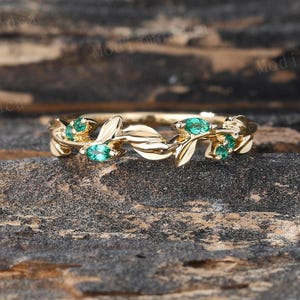 May include: A gold ring with a leaf and emerald design. The ring features small, oval-shaped emeralds set among delicate leaf-shaped details. The ring is displayed on a textured, dark brown surface.