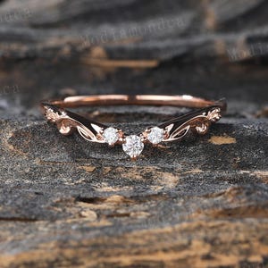 May include: A delicate rose gold wedding band with a curved design and three small diamonds. The band is set on a rustic wooden background.