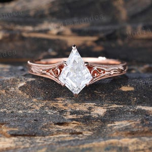 Peut inclure: Bague de fiançailles en or rose avec un diamant taille cerf-volant. La monture présente un motif de filigrane complexe. Le diamant est serti de griffes. La bague est présentée sur une surface texturée.