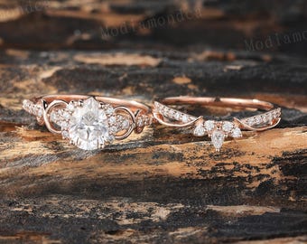 May include: A rose gold engagement ring set featuring a pear-shaped center stone and a curved wedding band. The engagement ring has a halo of smaller stones and intricate detailing. The rings are displayed on a textured wooden surface.