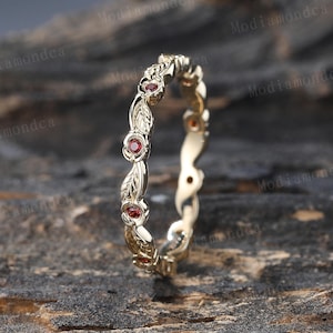 Puede incluir: Un delicado anillo de banda de oro con un diseño de hojas y pequeñas piedras preciosas rojas. El anillo está colocado sobre una superficie de madera.