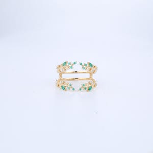 May include: A gold ring with two bands, decorated with small green gemstones and clear crystals. The ring has a leaf-like design, with the gemstones arranged to look like leaves. The ring is set against a white background.