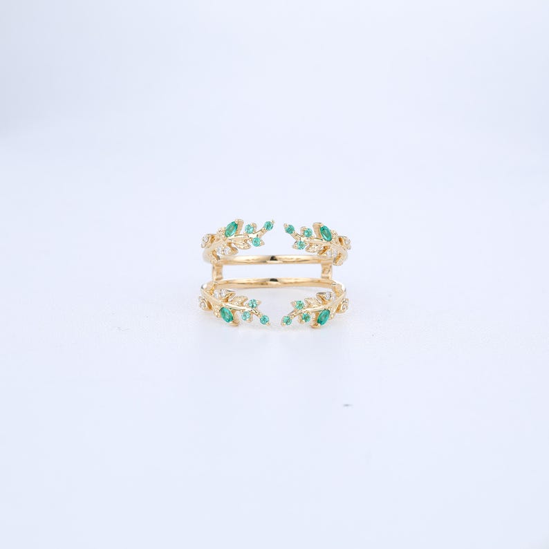 May include: A gold ring with two bands, decorated with small green gemstones and clear crystals. The ring has a leaf-like design, with the gemstones arranged to look like leaves. The ring is set against a white background.
