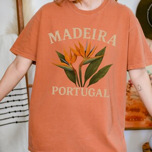Puede incluir: Camiseta naranja con las palabras "MADEIRA PORTUGAL" en una fuente marrón claro. La camiseta presenta un diseño floral con flores naranjas y azules y hojas verdes. La camiseta tiene mangas cortas.