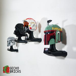 May include: Collection of Star Wars LEGO helmets displayed on black stands. Includes a silver helmet, a black Darth Vader helmet, a red and white X-wing pilot helmet, and a green and red Boba Fett helmet. The Gecko Bricks logo is in the bottom left corner.