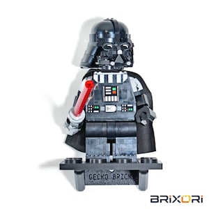May include: A black plastic construction toy figure of Darth Vader with a helmet, cape, and a red lightsaber. The figure stands on a black display base with the text GECKO BRICK printed on the front. The chest panel features small colored buttons.
