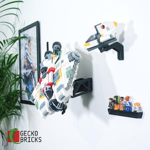 Könnte beinhalten: Weiße, rote und graue Lego-Raumschiffmodelle, die mit schwarzen Halterungen an einer weißen Wand befestigt sind. Ein gerahmtes Bild und ein kleines Regal mit Lego-Figuren sind ebenfalls zu sehen. Das Gecko Bricks-Logo befindet sich unten links.