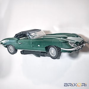 May include: A dark green model sports car constructed from building blocks, featuring a black convertible top, a clear windshield, silver-colored wire-style wheels, and a low-profile body design mounted on a black display bracket against a white background.