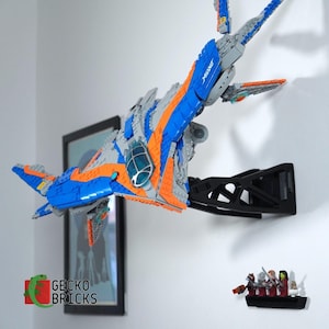 May include: A blue, orange, and gray Milano spaceship model made of interlocking plastic bricks, mounted on a black wall bracket. The model is displayed on a white wall, with a framed picture and a small display of figurines visible.