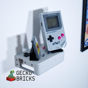May include: A gray Nintendo Game Boy console with a game cartridge and a stand. The Game Boy has a screen displaying a game, pink buttons, and a black directional pad. The text "Nintendo GAME BOY" is visible. The text "GECKO BRICKS" is also visible.