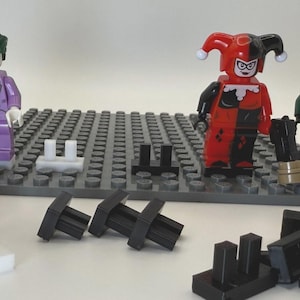 May include: A collection of Lego minifigures featuring characters from DC Comics and Star Wars. The Joker, Harley Quinn, and a clone trooper stand on a gray baseplate, with additional black and white Lego pieces scattered around.