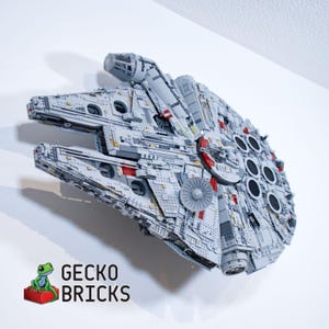 Op de afbeelding: Een gedetailleerd model van het Millennium Falcon ruimteschip, geconstrueerd uit grijze en rode bouwstenen. Het model is op een witte muur gemonteerd. De tekst "GECKO BRICKS" wordt in zwart en wit weergegeven.