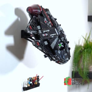 Könnte beinhalten: Schwarzes Lego-Modell eines Raumschiffs mit roten und grauen Details, an einer weißen Wand befestigt. Ein kleineres Lego-Modell mit Figuren und roten Lichtschwertern ist darunter montiert. Das Gecko Bricks-Logo befindet sich unten rechts.