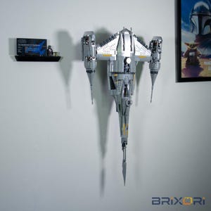 May include: A silver and grey LEGO Star Wars N-1 Starfighter model mounted vertically on a wall. The spacecraft features a long, pointed nose, two side engines, and a cockpit. A small display plaque and two minifigures sit on a nearby shelf.