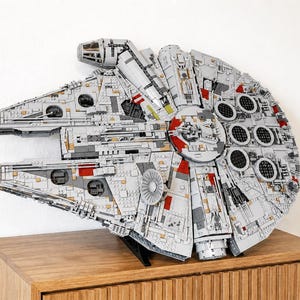 Puede incluir: Un modelo detallado de la nave espacial Halcón Milenario, construido con bloques de construcción entrelazados. El modelo presenta una combinación de colores gris y blanco con detalles en rojo y negro. Se exhibe sobre un mueble de madera contra una pared blanca.