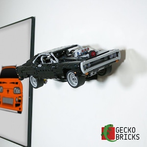 Könnte beinhalten: Ein schwarzes Modell eines Muscle-Cars aus Bausteinen, das horizontal an einer Wand montiert ist. Das Fahrzeug verfügt über einen freiliegenden Motorblock mit roten und blauen Details, silberne Räder und ein detailliertes Fahrgestell. Text Gecko Bricks.