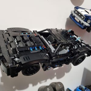 May include: A black Lego Batmobile model with intricate details, including a detailed engine and wheels. The model is displayed with a blue and white Lego car in the background. The Batmobile has blue and red accents.
