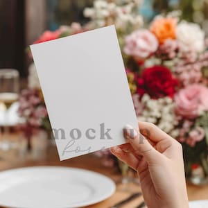 May include: A blank white card held in a hand, with the text "mock for" visible. The background features a blurred floral arrangement with pink, red, and orange flowers, and a white plate.