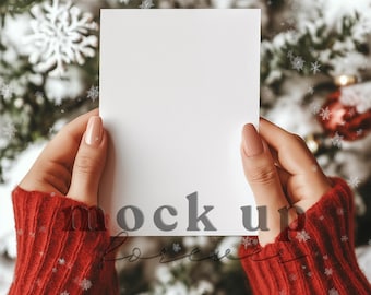 May include: A blank white card held by hands wearing a red sweater. The card is in front of a blurred Christmas tree with red ornaments and snowflakes. The words "mock up forever" are at the bottom of the card.
