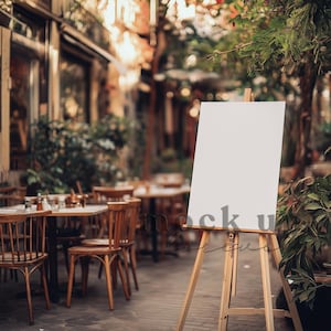 May include: A blank white sign on an easel stands in front of a cafe with tables and chairs. The cafe is located on a cobblestone street with trees and greenery in the background.
