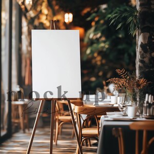 May include: A white canvas on an easel in a restaurant setting. The canvas is blank and ready for a sign or artwork. The restaurant has wooden chairs and tables with a white tablecloth.