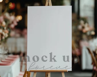May include: A white easel with a blank sign, perfect for displaying wedding information or a welcome message. The easel is set up in a wedding reception venue with tables and chairs in the background.