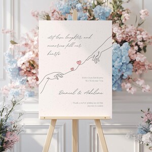 May include: A white sign with a hand-drawn illustration of two hands reaching towards each other with hearts connecting them. The text reads "Let love, laughter, and memories fill our hearts. With grateful hearts, we welcome you. Daniel & Adeline. Thank you for joining us on this joyous occasion."