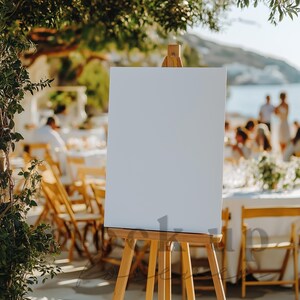 May include: A white easel with a blank canvas, set up outdoors at a wedding reception. The easel is in front of a table with chairs and guests in the background.