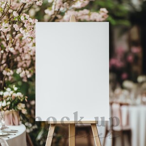 May include: A white blank sign on an easel in front of a blurred background of pink flowers and a table setting.