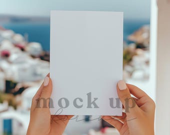 May include: A blank white card held by two hands against a blurred background of a white building and blue water.
