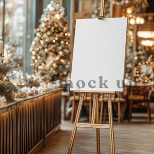May include: A wooden easel with a blank white canvas, perfect for displaying artwork or signage. The word "mock up" is printed on the canvas. The easel is set in a festive, decorated space with a Christmas tree in the background.
