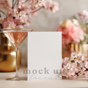 May include: A martini glass with a gold rim filled with a pink liquid stands next to a white card with the text "mock up forever". Pink flowers and a gold vase are in the background.