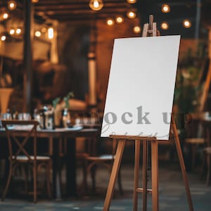 Op de afbeelding: Een leeg wit bord op een ezel in een rustieke omgeving. Het bord staat voor een tafel met stoelen en lichtslingers.
