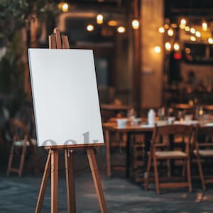Op de afbeelding: Een houten ezel met een leeg wit doek, staand voor een restaurant met lichtslingers en houten tafels en stoelen.