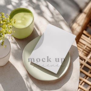 Puede incluir: Una tarjeta blanca en blanco con el texto "mock up forever" descansa sobre un plato verde claro. Una taza verde claro de té matcha y un pequeño jarrón con flores amarillas también están sobre la mesa. La escena está iluminada por la luz del sol.