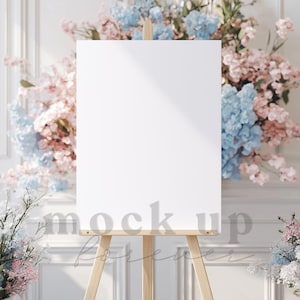 Pode incluir: Uma tela branca em um cavalete com uma moldura de madeira, cercada por flores rosa e azuis. A tela está em branco e pronta para uma placa ou uma obra de arte.