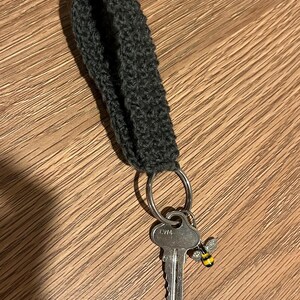 Puede incluir: Un llavero de pulsera de tela tejida a ganchillo en color gris oscuro con una anilla de metal plateado. Enganchada a la anilla hay una llave de metal plateado con el texto CW4 grabado en la cabeza y un pequeño dije de abeja a rayas amarillas y negras.